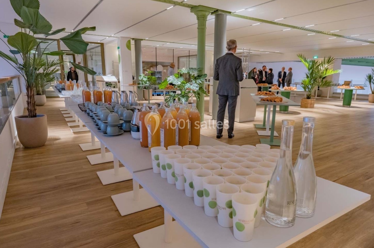 Buffet avec jus d'orange, tasses, bouteilles d'eau et viennoiseries dans une salle lumineuse avec plantes et invités en…