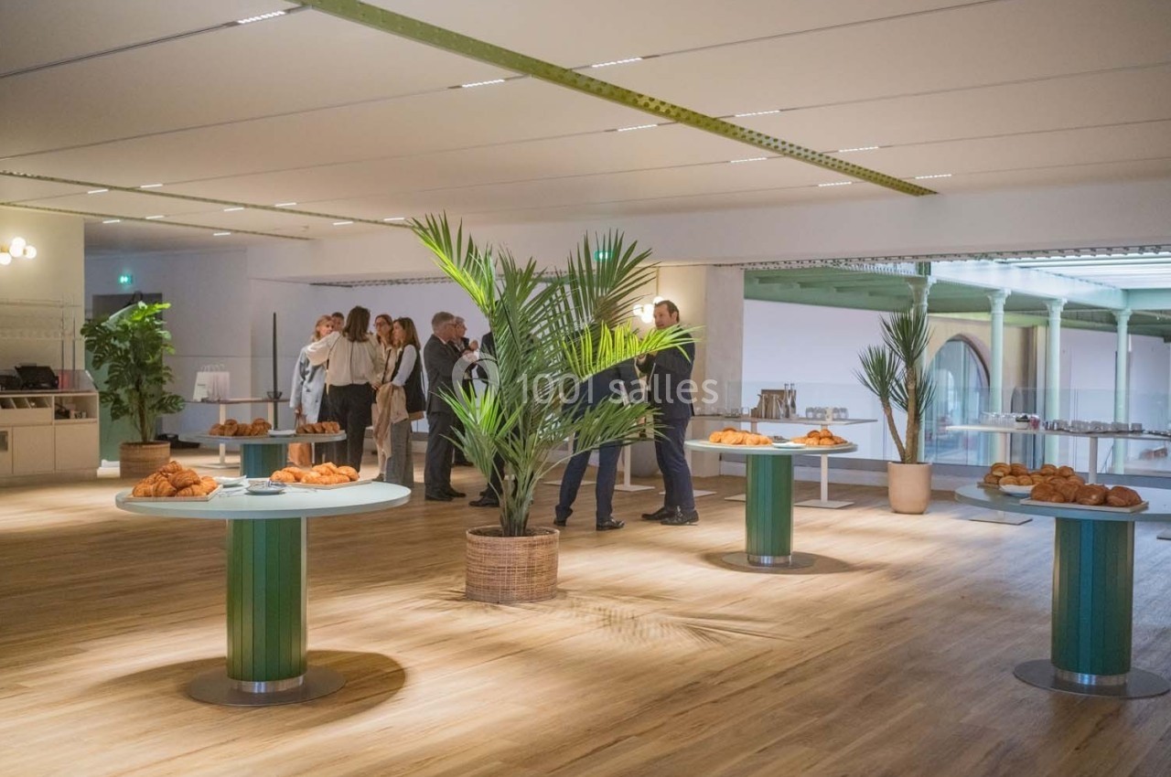 Salle lumineuse avec parquet, tables garnies de viennoiseries et groupe de personnes discutant en arrière-plan.