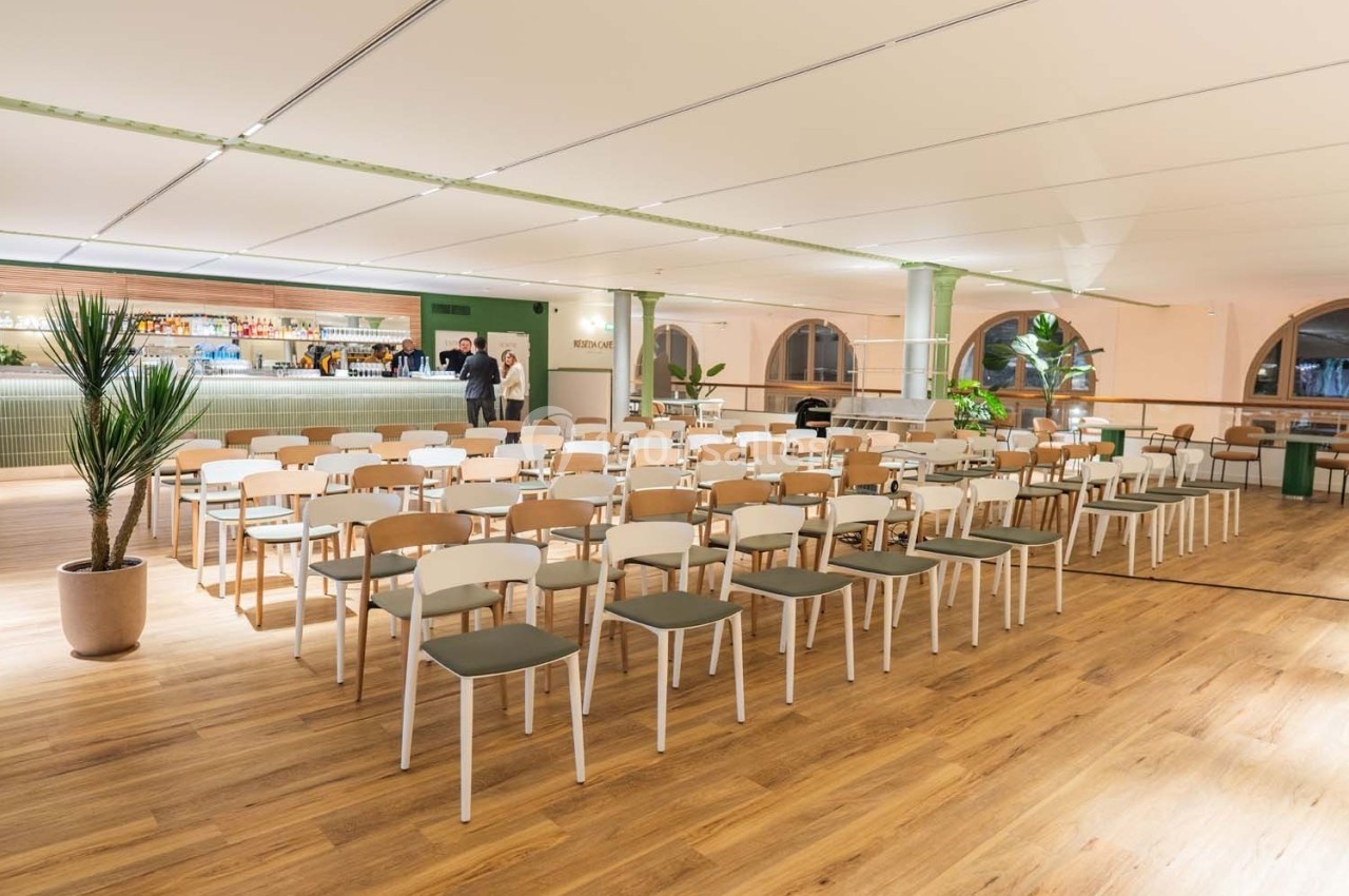 Salle lumineuse avec des rangées de chaises alignées face à un bar, sol en bois et plantes décoratives.