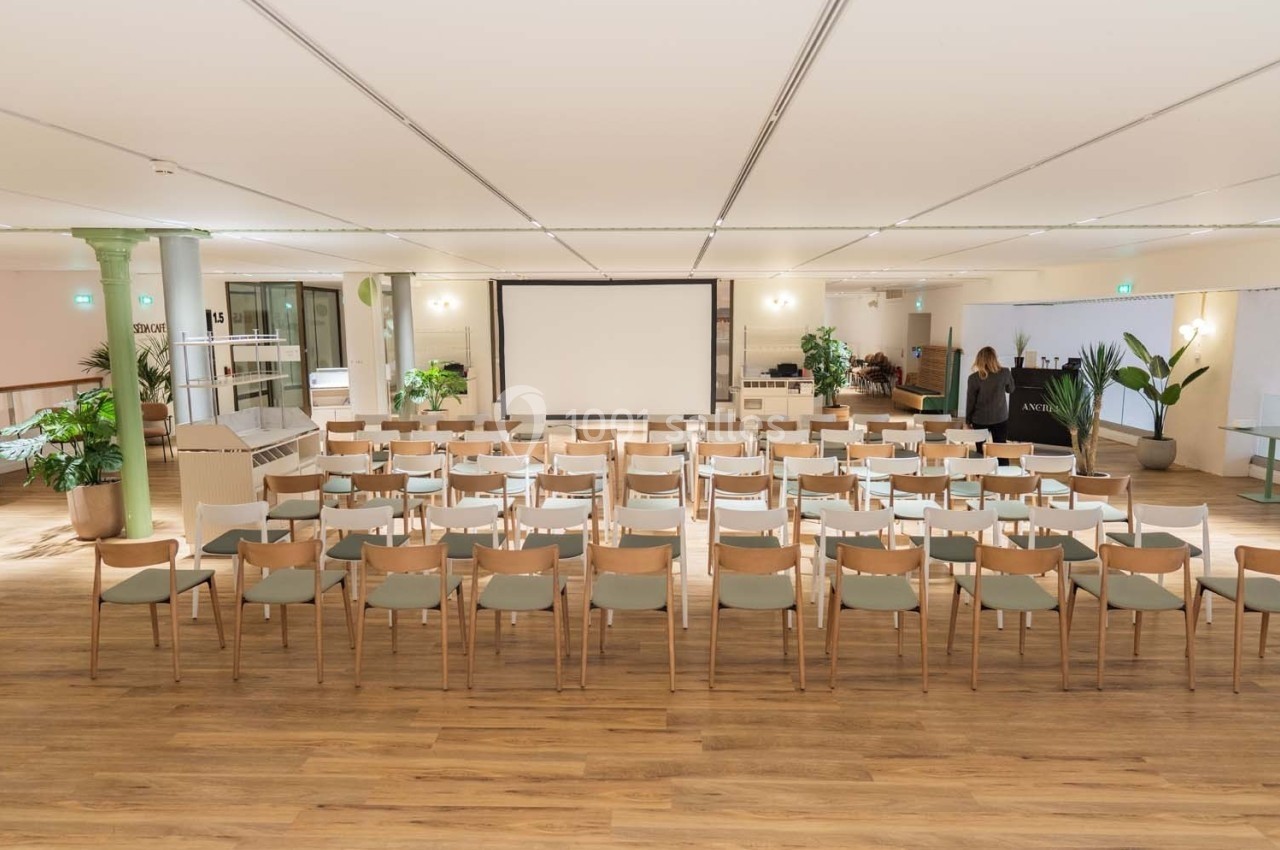 Salle lumineuse avec rangées de chaises en bois disposées face à un écran de projection, entourée de plantes.