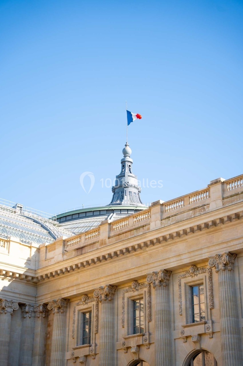 Dôme et façade d'un bâtiment historique avec un drapeau français flottant sous un ciel bleu.