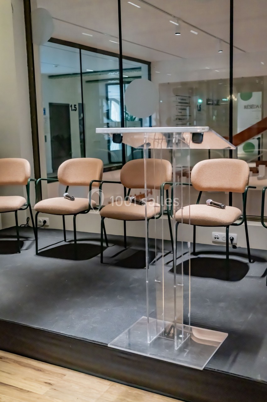 Un pupitre transparent sur une scène, entouré de chaises vides dans une salle de conférence moderne.