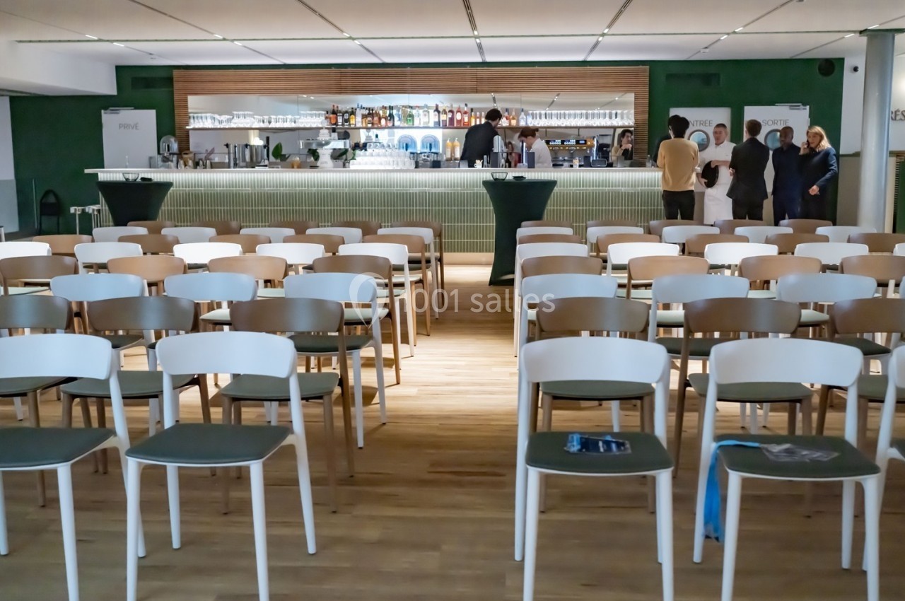 Salle avec des rangées de chaises vides face à un bar, quelques personnes discutant en arrière-plan.