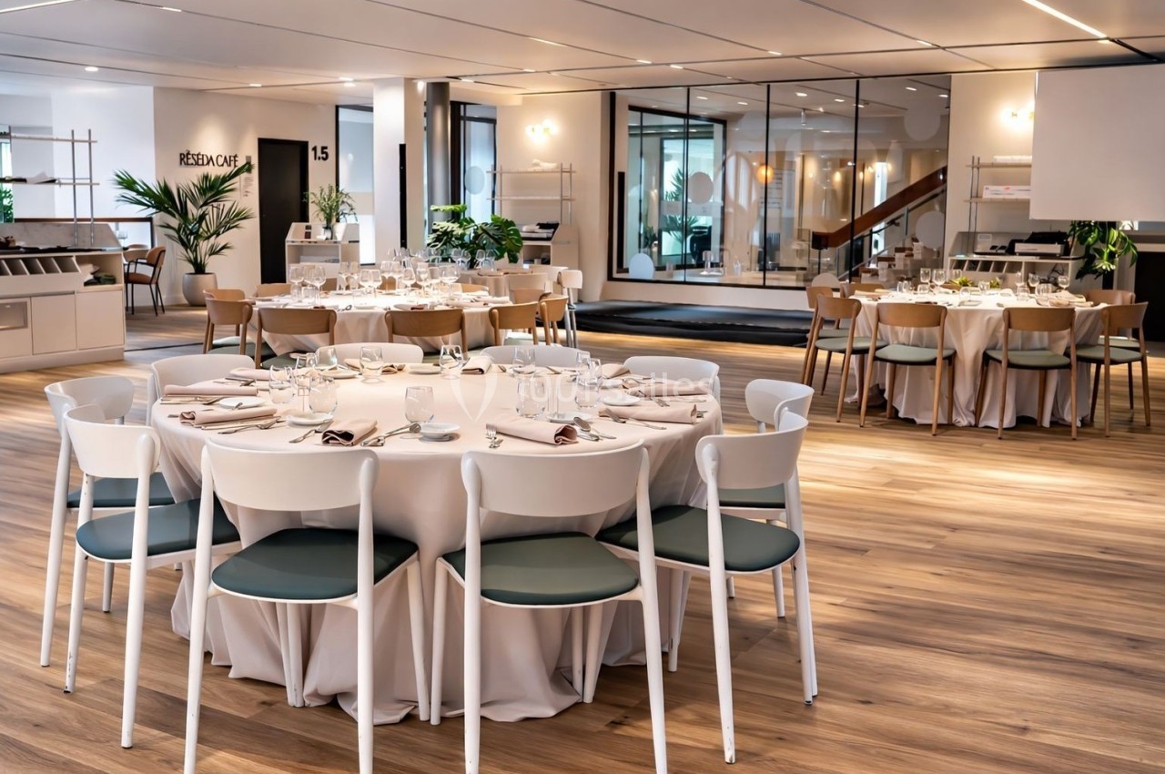 Salle lumineuse avec tables rondes dressées pour un repas, chaises blanches et décor moderne en arrière-plan.