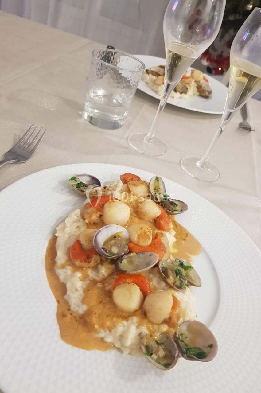 Assiette de risotto crémeux garni de noix de Saint-Jacques, palourdes et sauce, servie sur une table dressée.