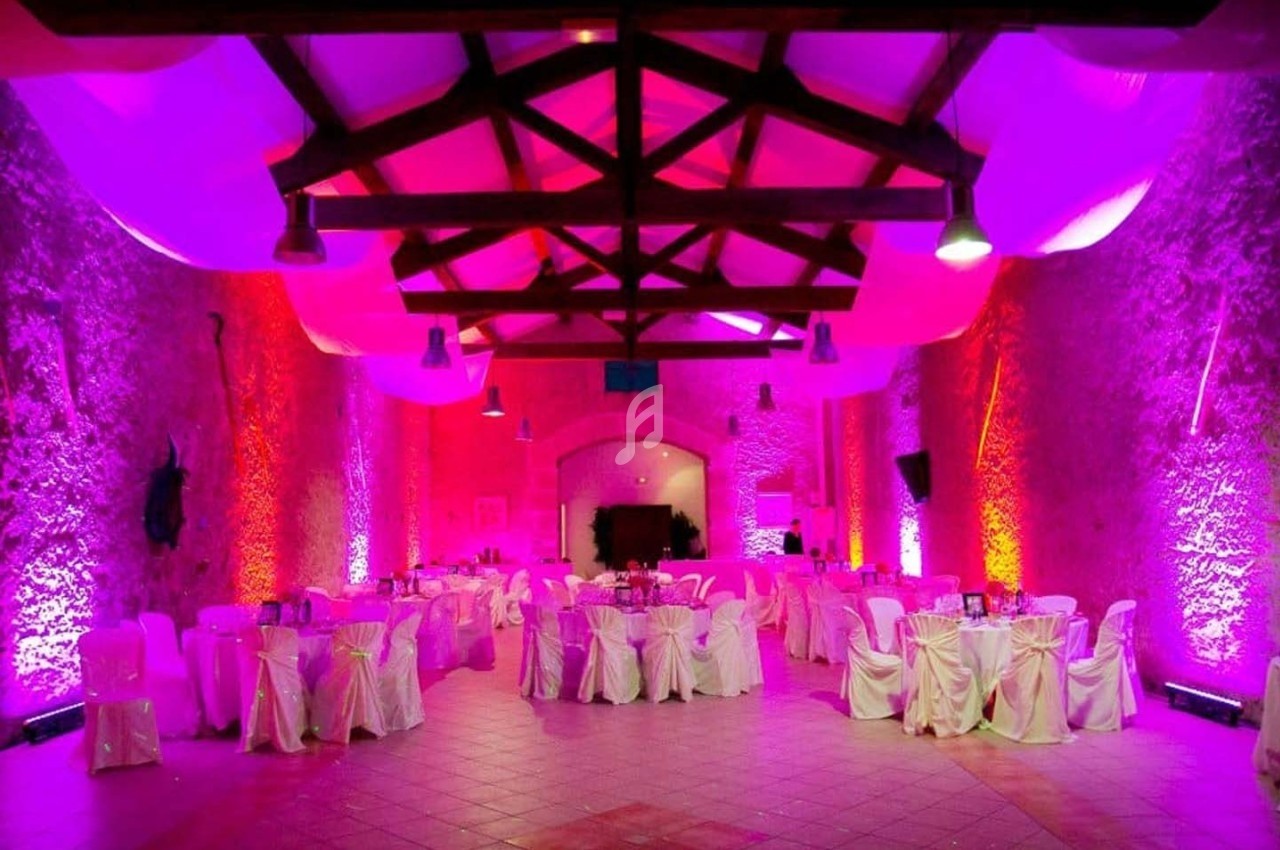 Salle de réception avec murs en pierre, éclairage coloré et tables rondes décorées de nappes blanches.
