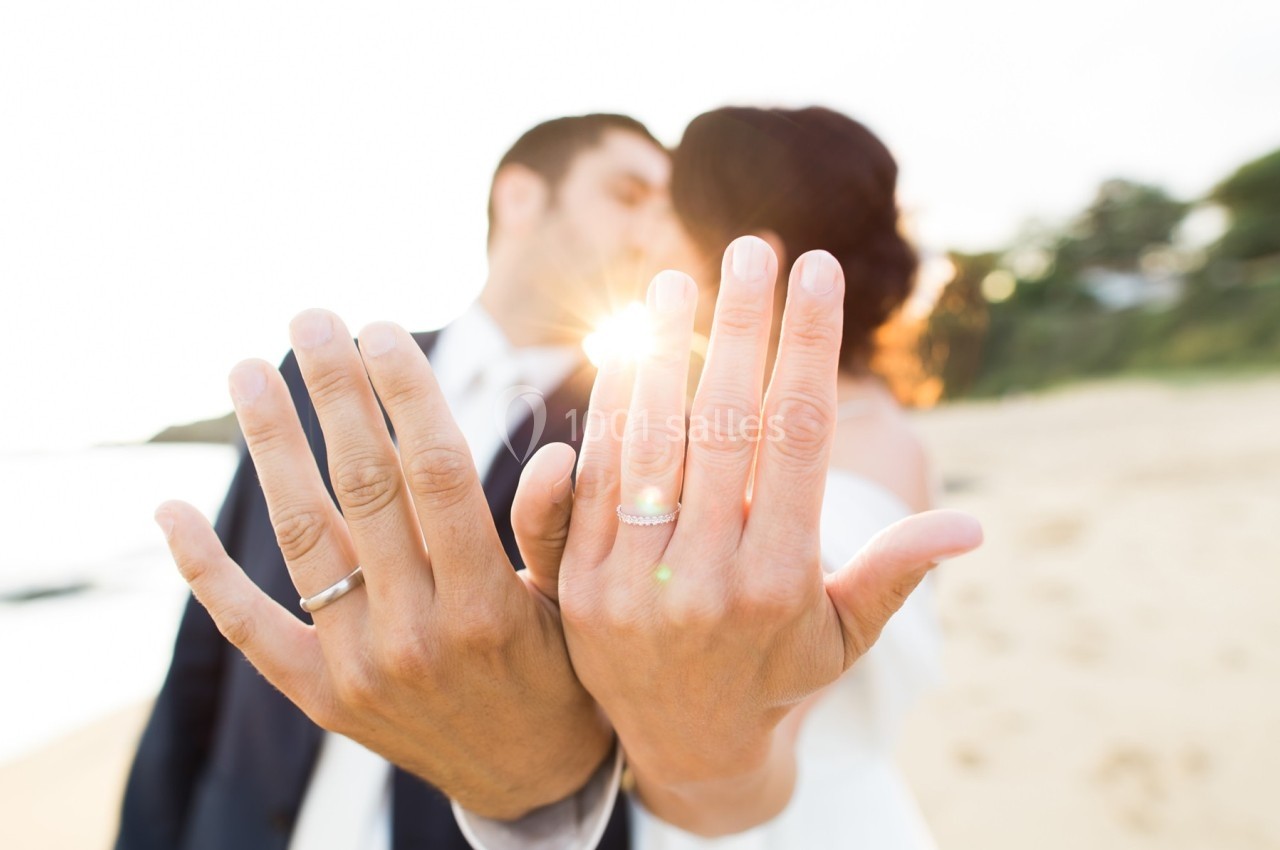 Un couple montre leurs alliances au premier plan sur une plage ensoleillée, flou en arrière-plan.