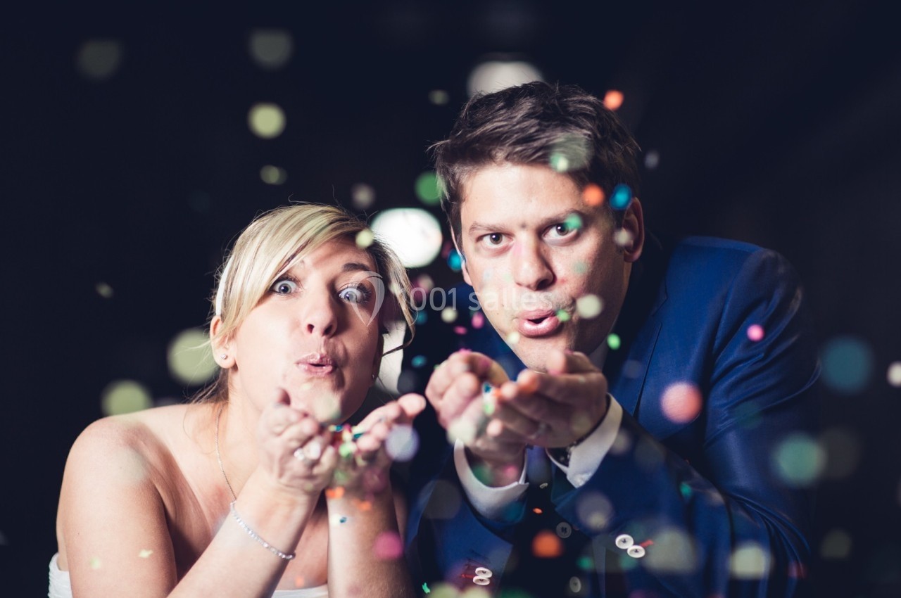 Un couple souffle des confettis colorés vers l'objectif dans un environnement sombre.