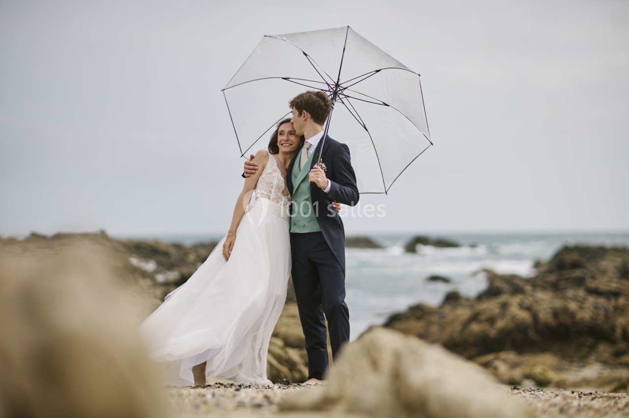 Un couple souriant sous un parapluie transparent sur une plage rocheuse par temps nuageux.
