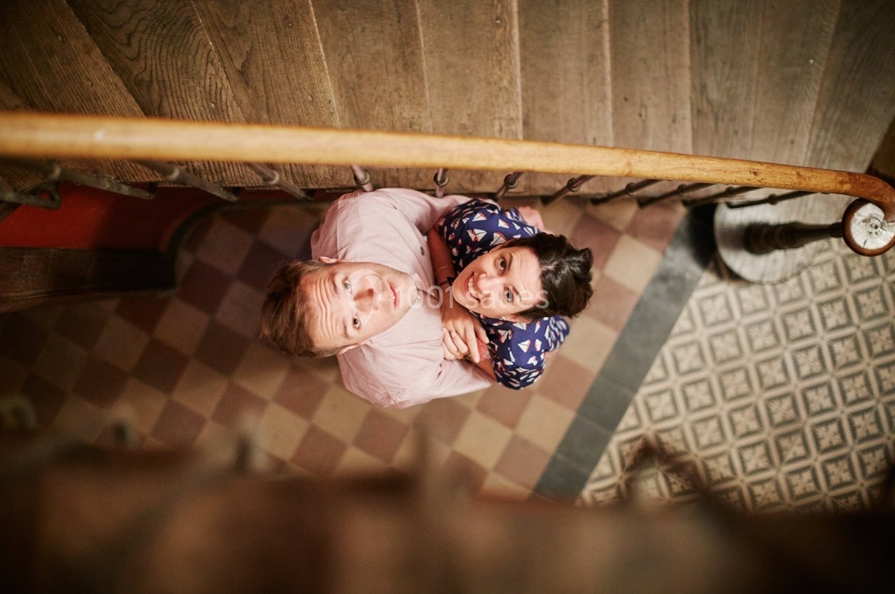 Vue plongeante d'un homme et d'une femme regardant vers le haut, debout sur un sol carrelé près d'un escalier en bois.