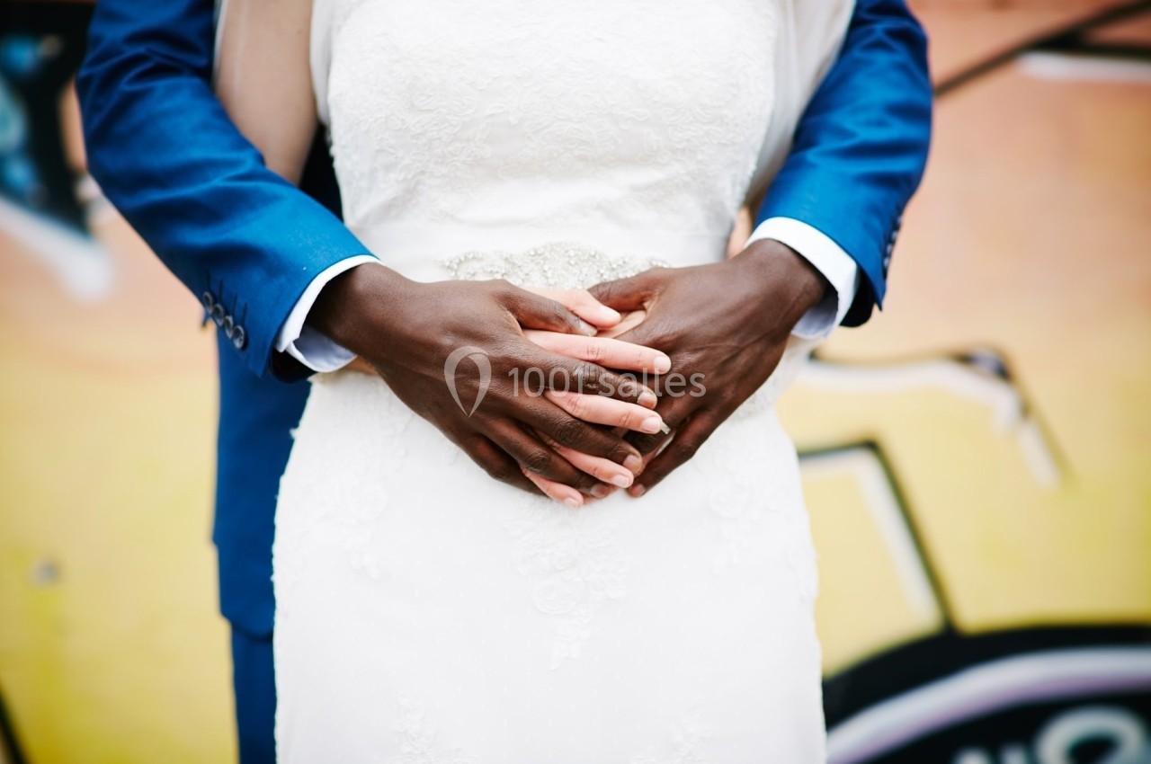 Un couple en tenue de mariage, l'homme en costume bleu tenant la taille de la femme en robe blanche devant un mur coloré.