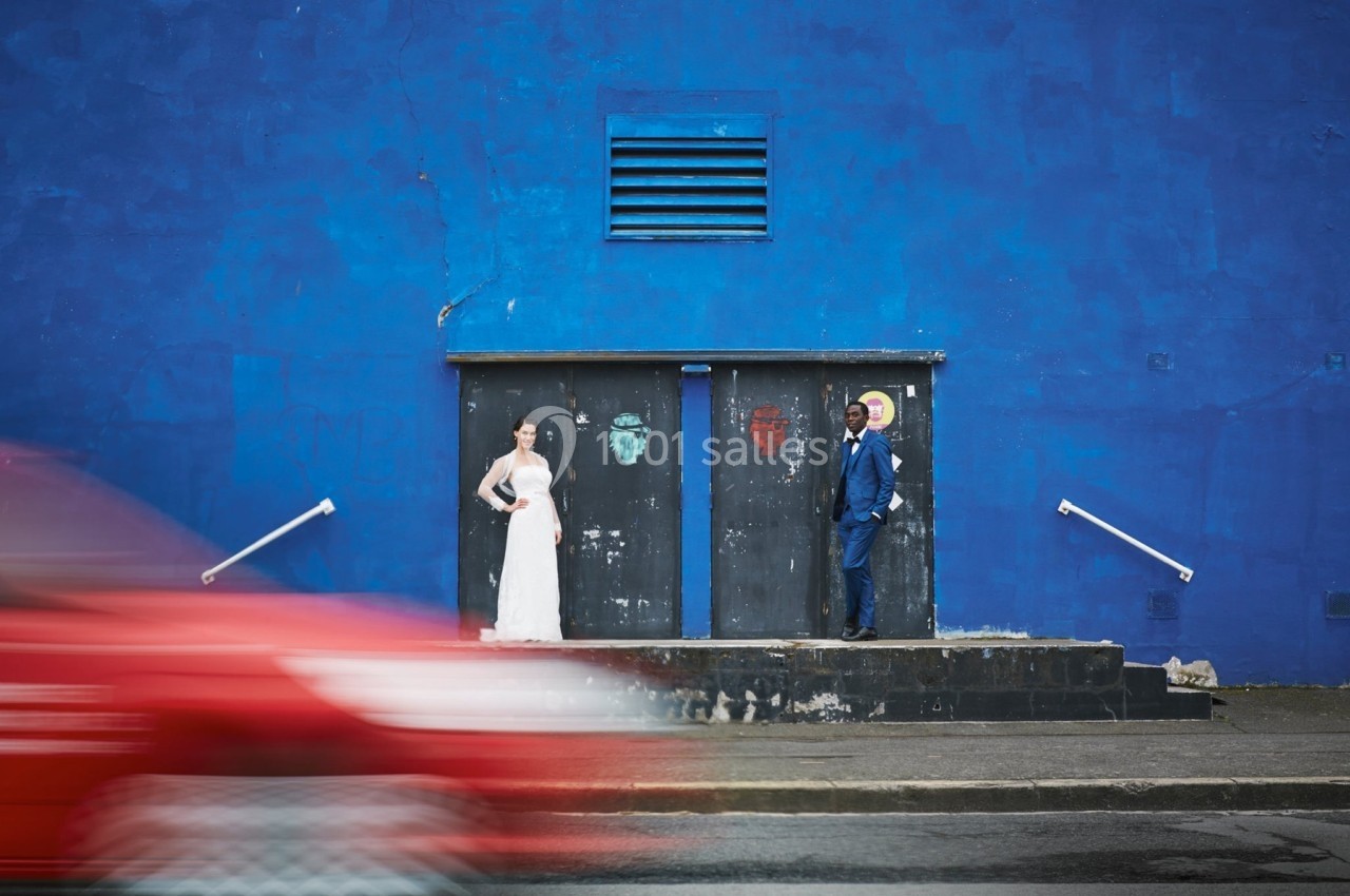 Un couple en tenue de mariage pose devant un mur bleu, avec une voiture rouge floue passant au premier plan.