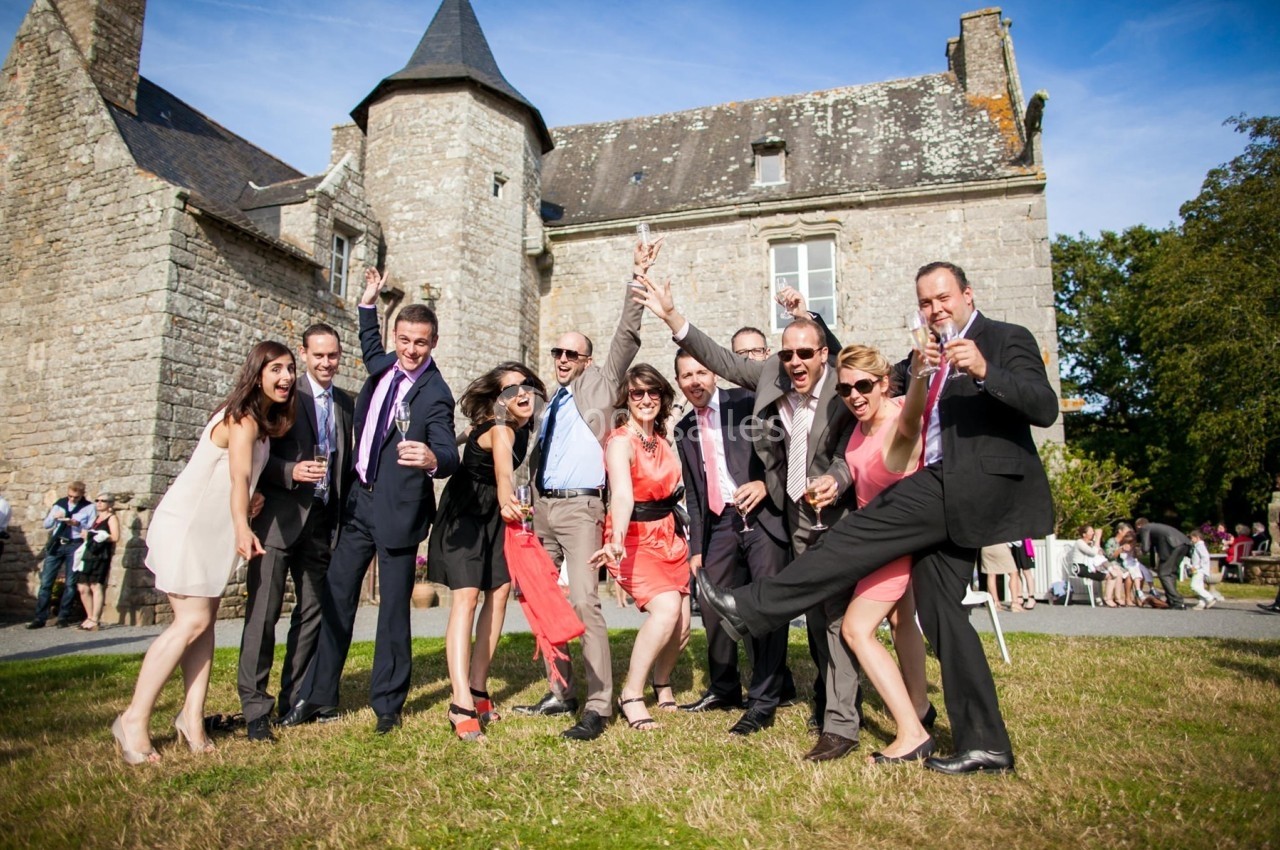 Un groupe de personnes bien habillées pose joyeusement devant un bâtiment en pierre sous un ciel dégagé.