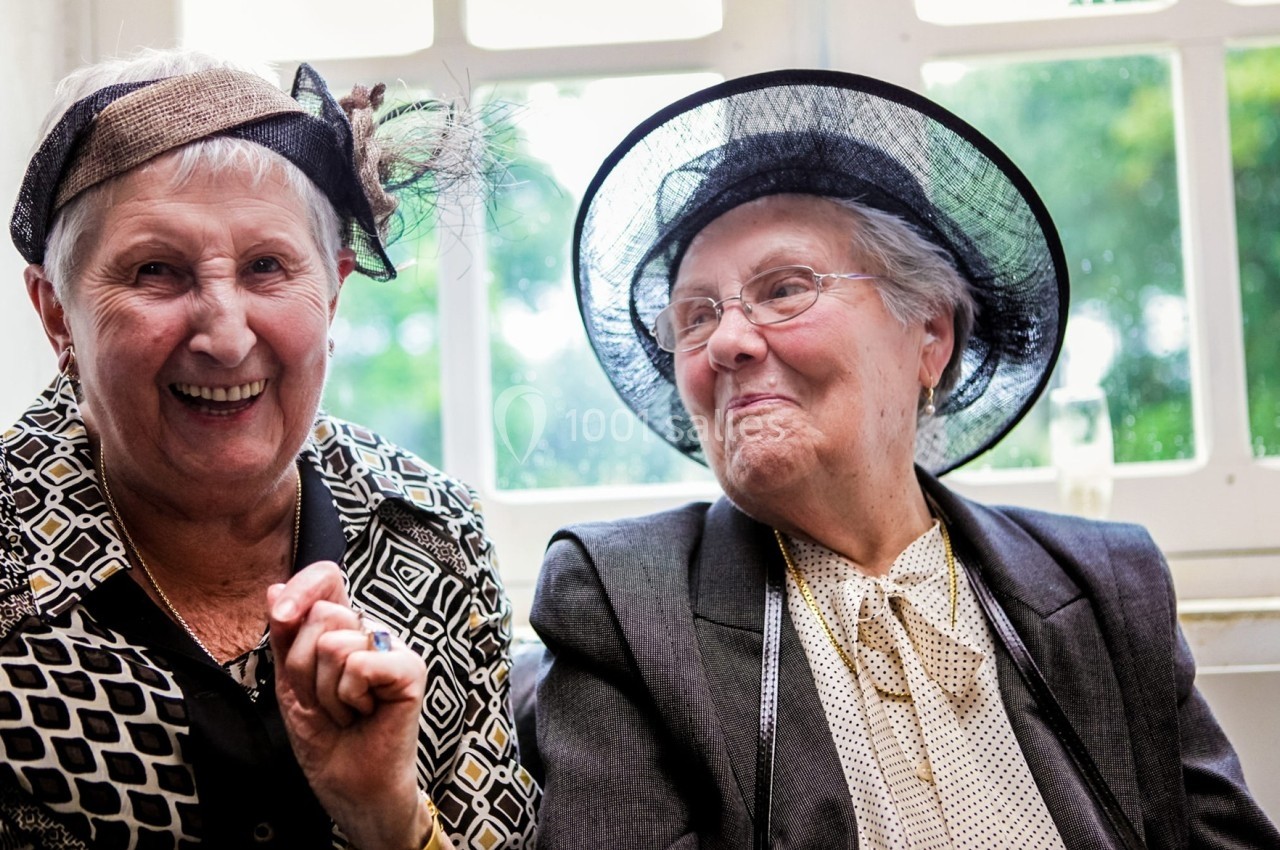 Deux femmes âgées souriantes portant des chapeaux élégants, assises près d'une fenêtre lumineuse.