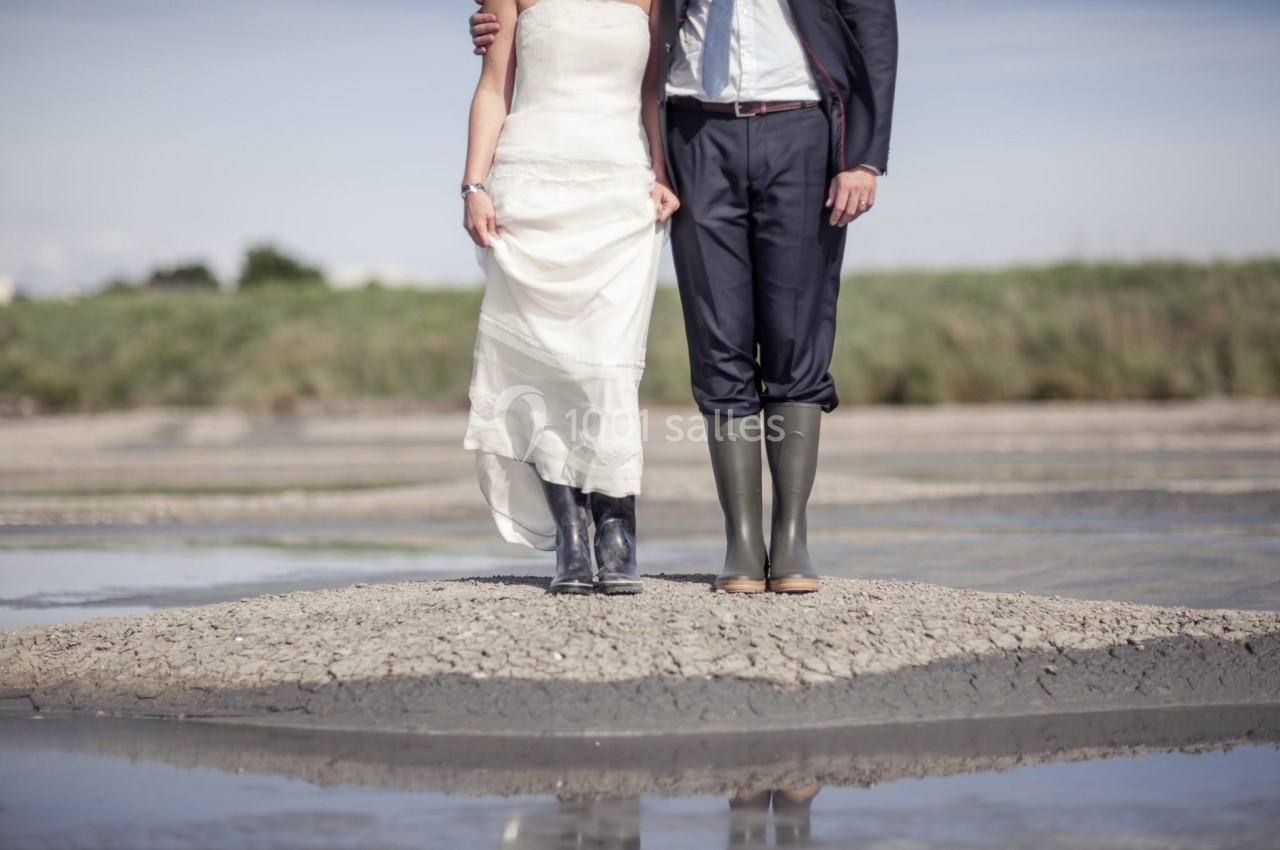 Un couple en tenue de mariage porte des bottes en caoutchouc, debout sur un sol sec entouré d'eau.