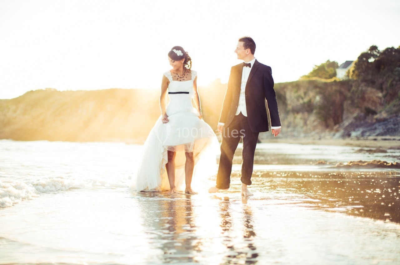 Un couple en tenue de mariage marche pieds nus sur une plage au coucher du soleil.