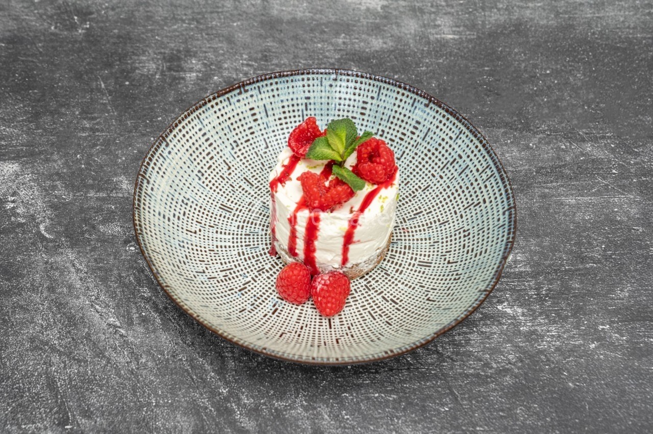 Dessert à base de fromage blanc, garni de framboises fraîches, coulis rouge et feuille de menthe, sur une assiette texturée.