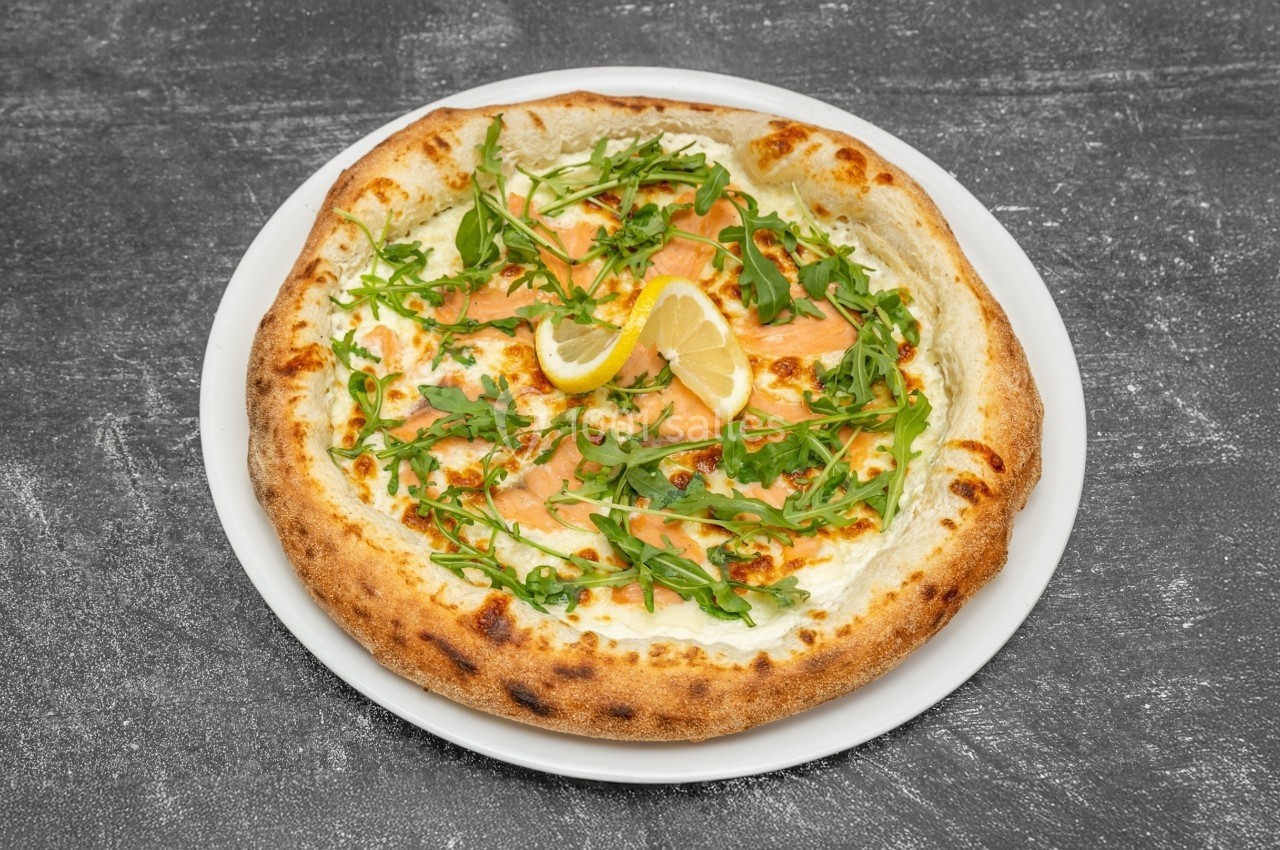 Pizza garnie de saumon fumé, roquette, citron et fromage, servie sur une assiette blanche sur fond sombre.