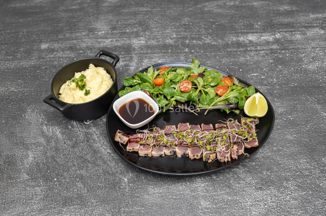 Assiette avec tataki de thon, salade verte, sauce soja, citron vert et purée servie dans une petite casserole noire.
