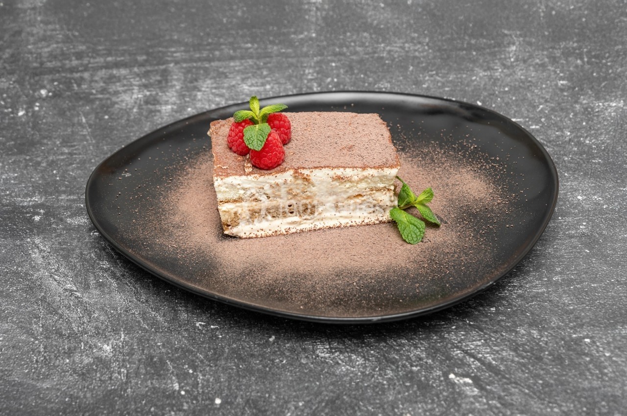 Part de tiramisu décorée de framboises et de feuilles de menthe, servie sur une assiette noire.