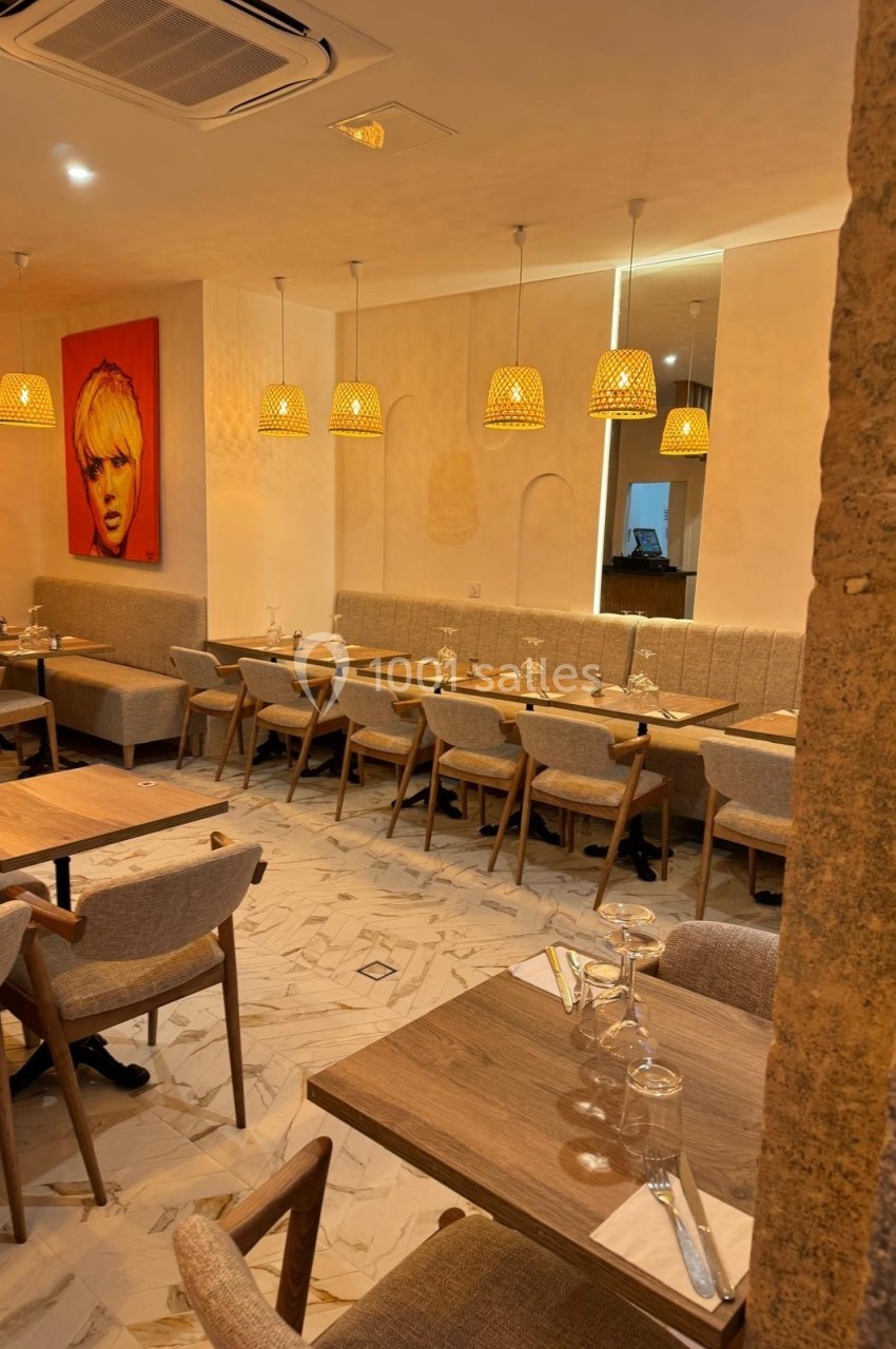 Salle de restaurant chaleureuse avec tables en bois, chaises rembourrées, luminaires suspendus et tableau mural coloré.
