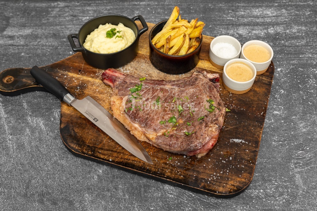 Côte de bœuf grillée servie sur une planche avec purée, frites, sauces et couteau.