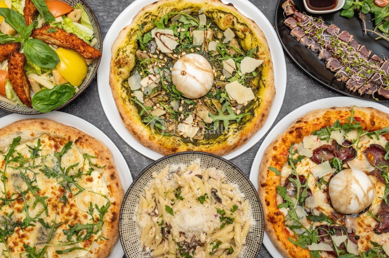 Assortiment de pizzas, pâtes et plats garnis de légumes et viandes, présenté sur une table vue de dessus.