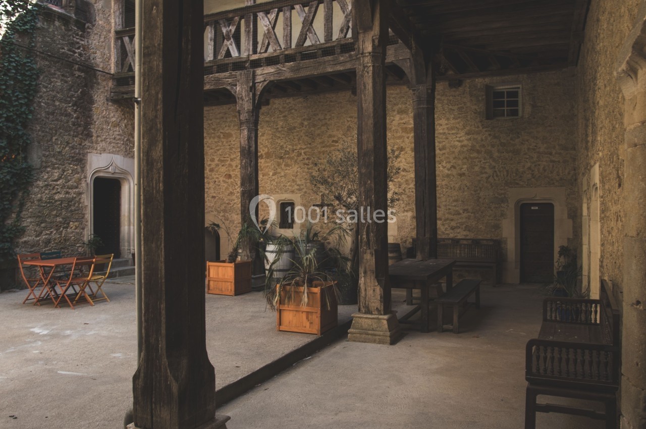 Cour intérieure avec murs en pierre, poutres en bois, tables, chaises orange et plantes en pots.