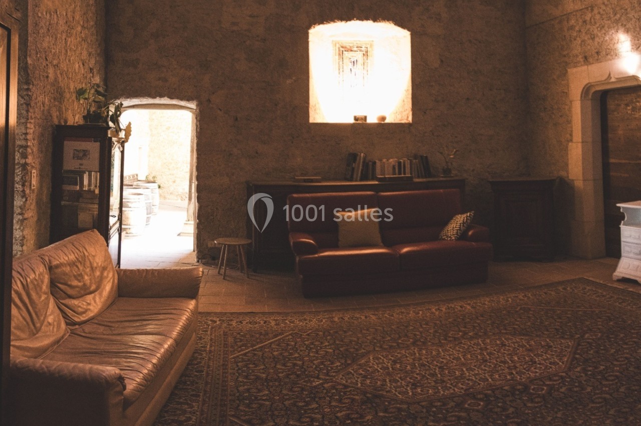 Salon rustique avec murs en pierre, canapés en cuir, tapis ancien et lumière naturelle entrant par une porte ouverte.