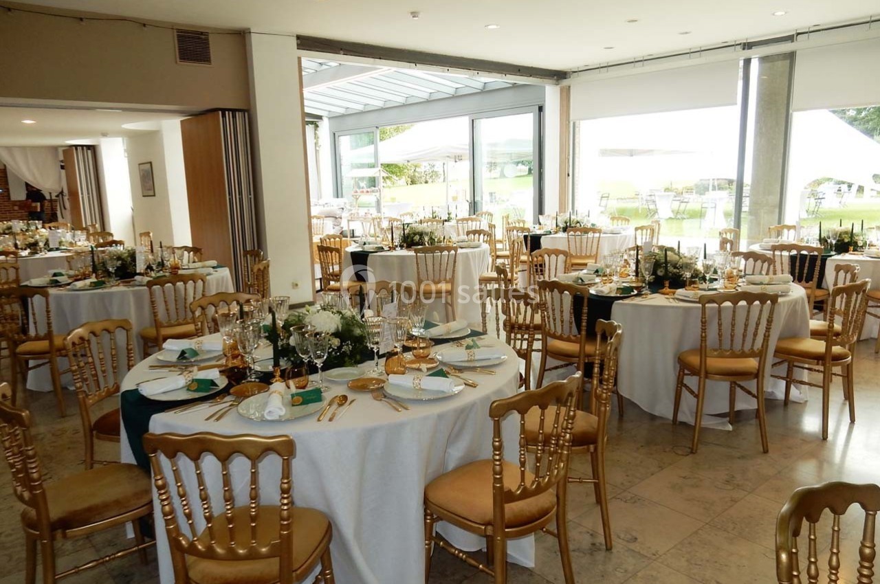 Salle de réception décorée avec des tables rondes dressées, chaises dorées et arrangements floraux élégants.