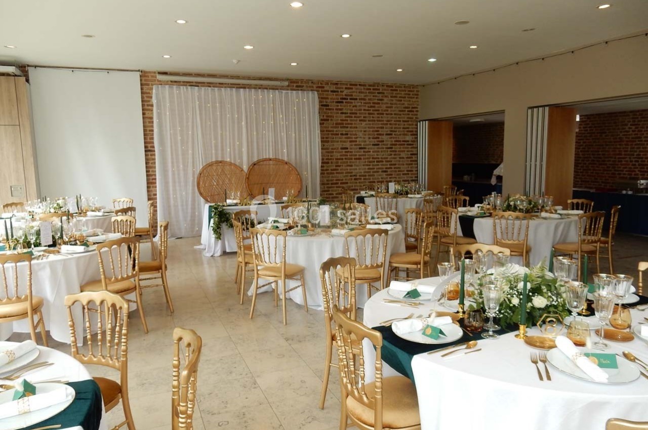 Salle de réception décorée avec des tables rondes, nappes blanches, chaises dorées et arrangements floraux élégants.