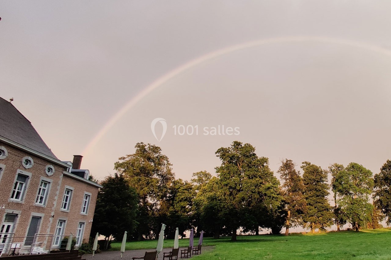 Un arc-en-ciel traverse un ciel clair au-dessus d'un bâtiment en briques et d'un parc verdoyant.