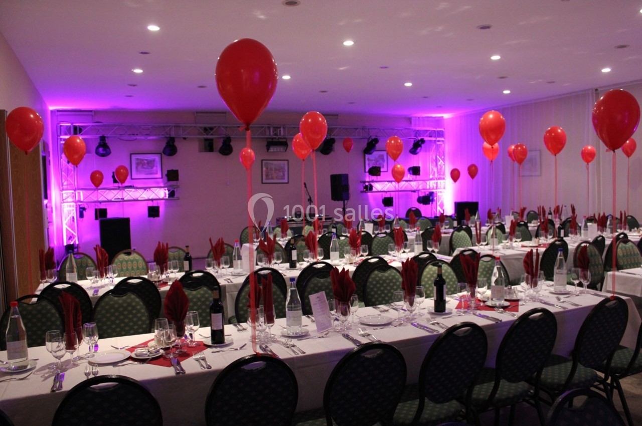 Salle décorée pour un événement avec tables dressées, ballons rouges flottants et éclairage violet.