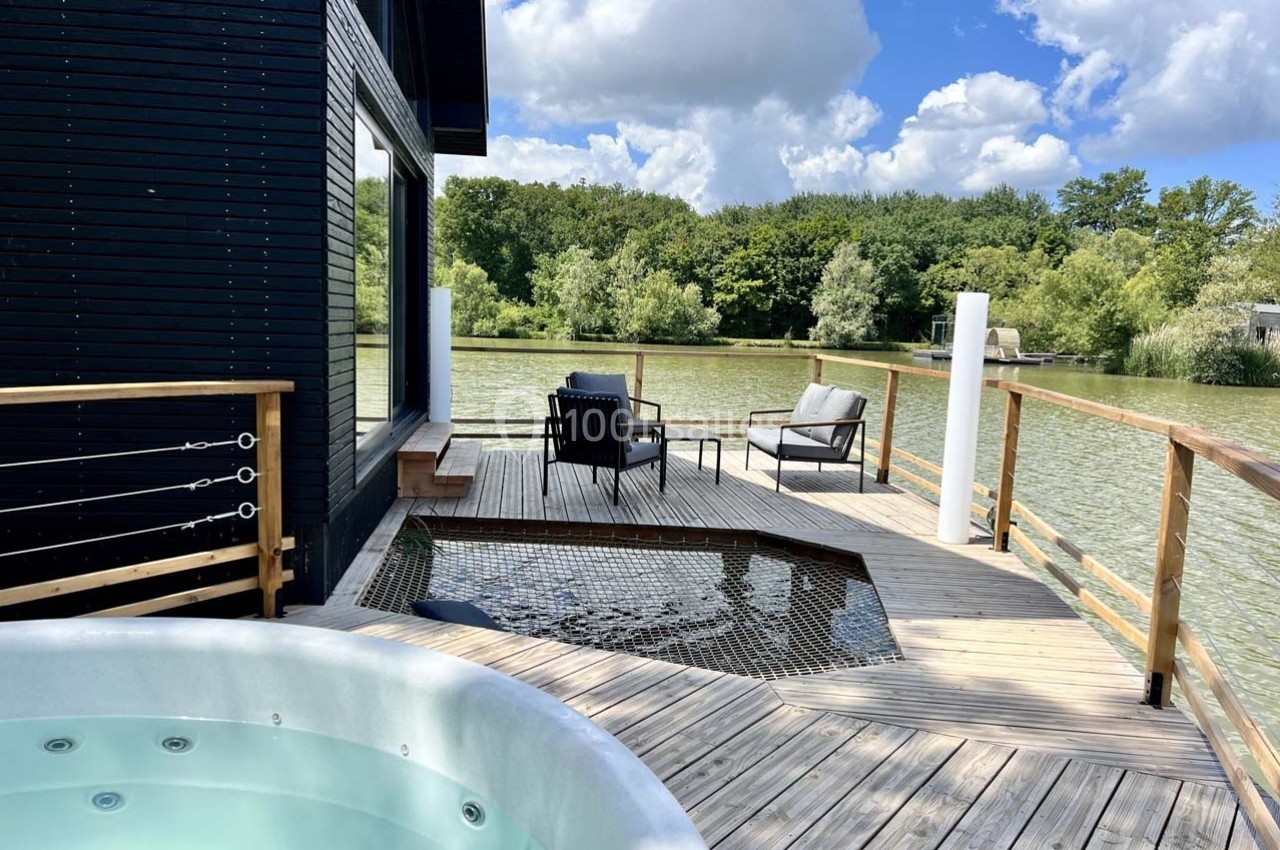 Terrasse en bois avec jacuzzi, filet suspendu, fauteuils et vue sur un lac entouré de végétation.