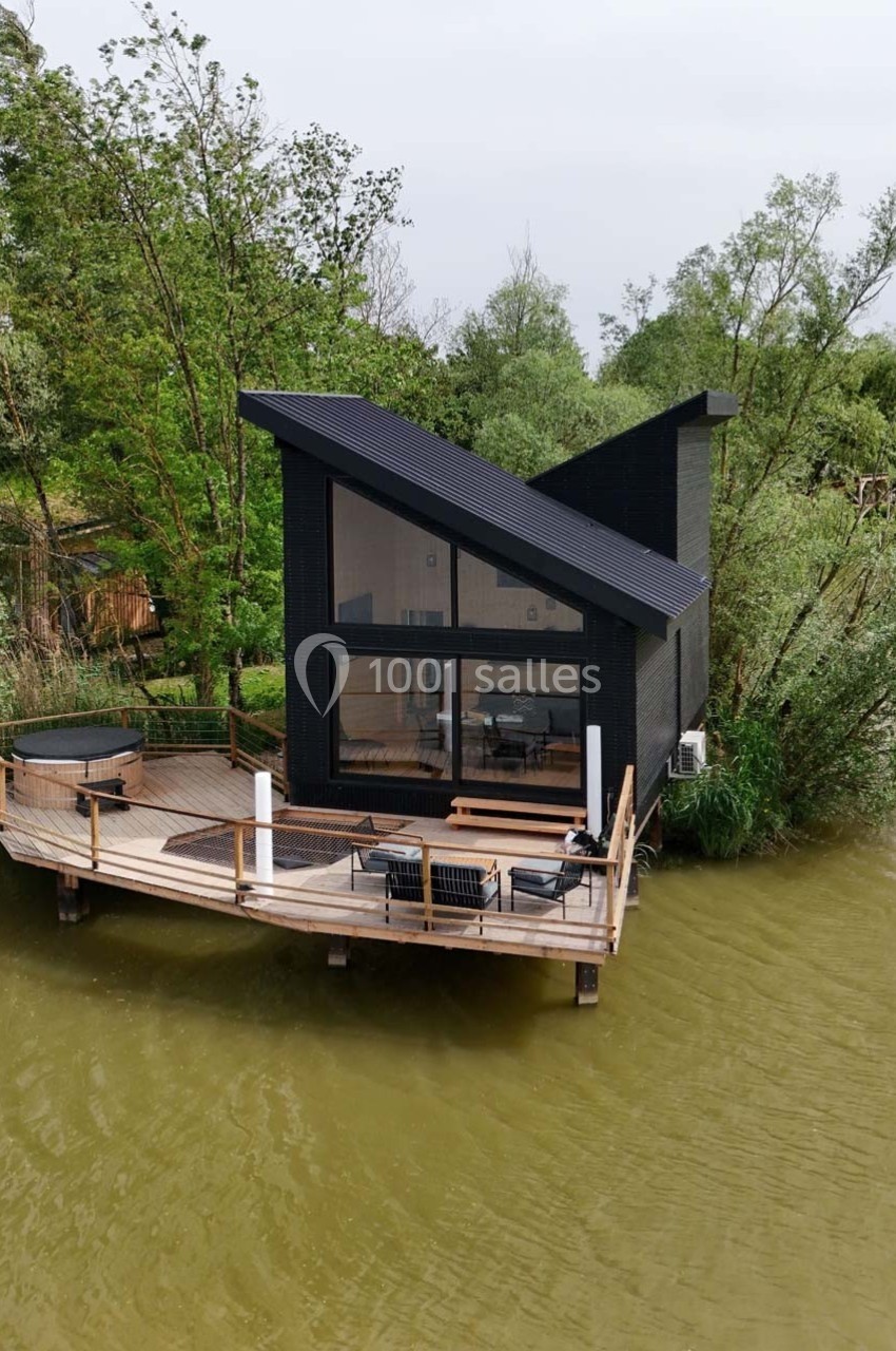 Cabane moderne en bois noir avec terrasse sur pilotis au bord d'un plan d'eau, entourée de végétation.