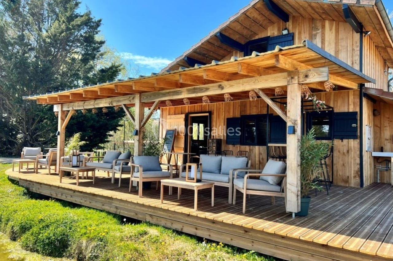 Terrasse en bois avec mobilier d'extérieur devant une maison en bois, entourée de végétation et d'un plan d'eau.