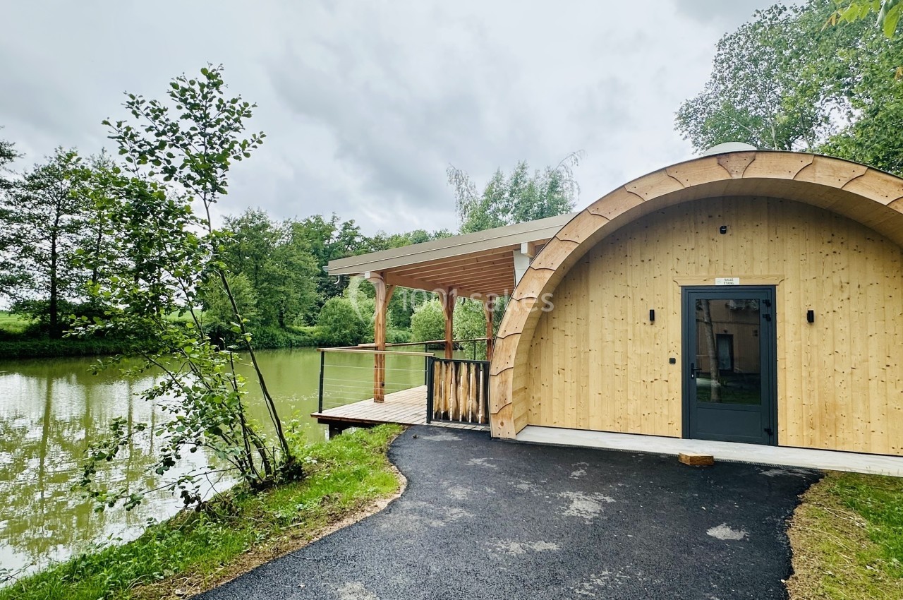 Cabane en bois arrondie avec terrasse couverte, située au bord d'un étang entouré de verdure.