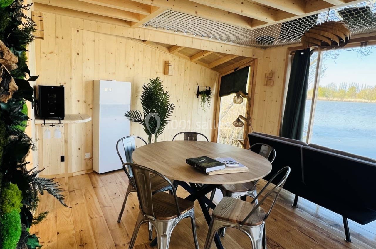 Salle à manger en bois avec table ronde, chaises métalliques, canapé, plantes et vue sur un lac par une baie vitrée.