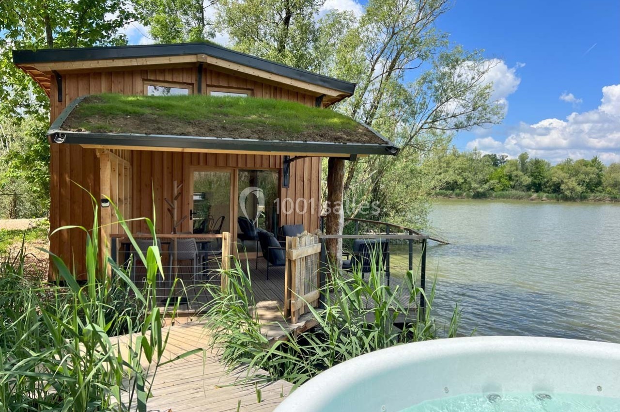 Cabane en bois avec toit végétalisé au bord d'un lac, entourée de végétation, avec un jacuzzi au premier plan.