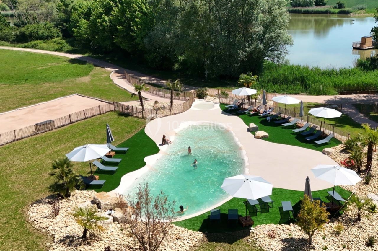 Vue aérienne d'une piscine extérieure entourée de transats, parasols, pelouse artificielle et végétation, près d'un lac.