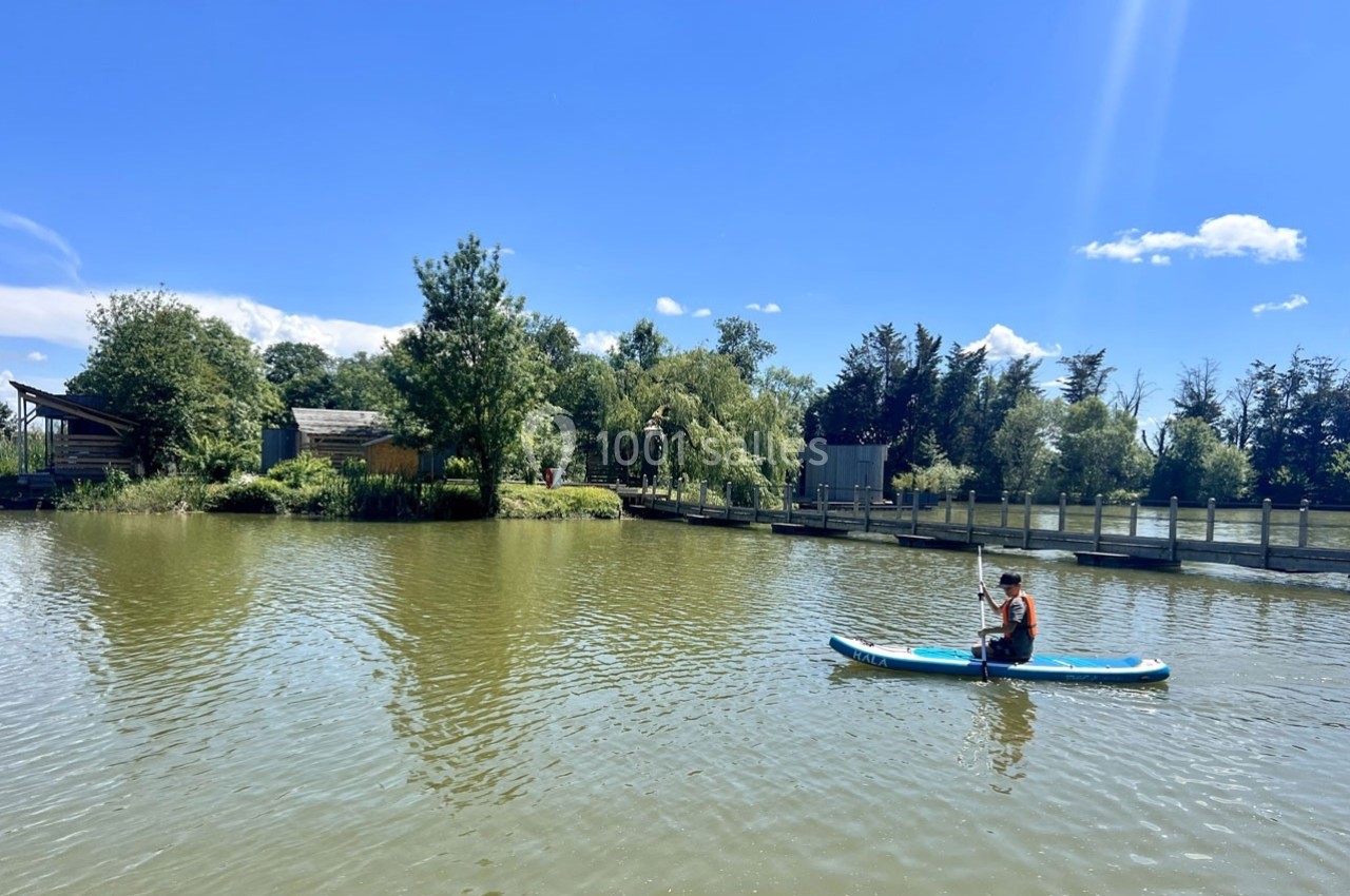 Une personne fait du paddle sur un lac entouré de végétation et de petites structures en bois sous un ciel bleu.