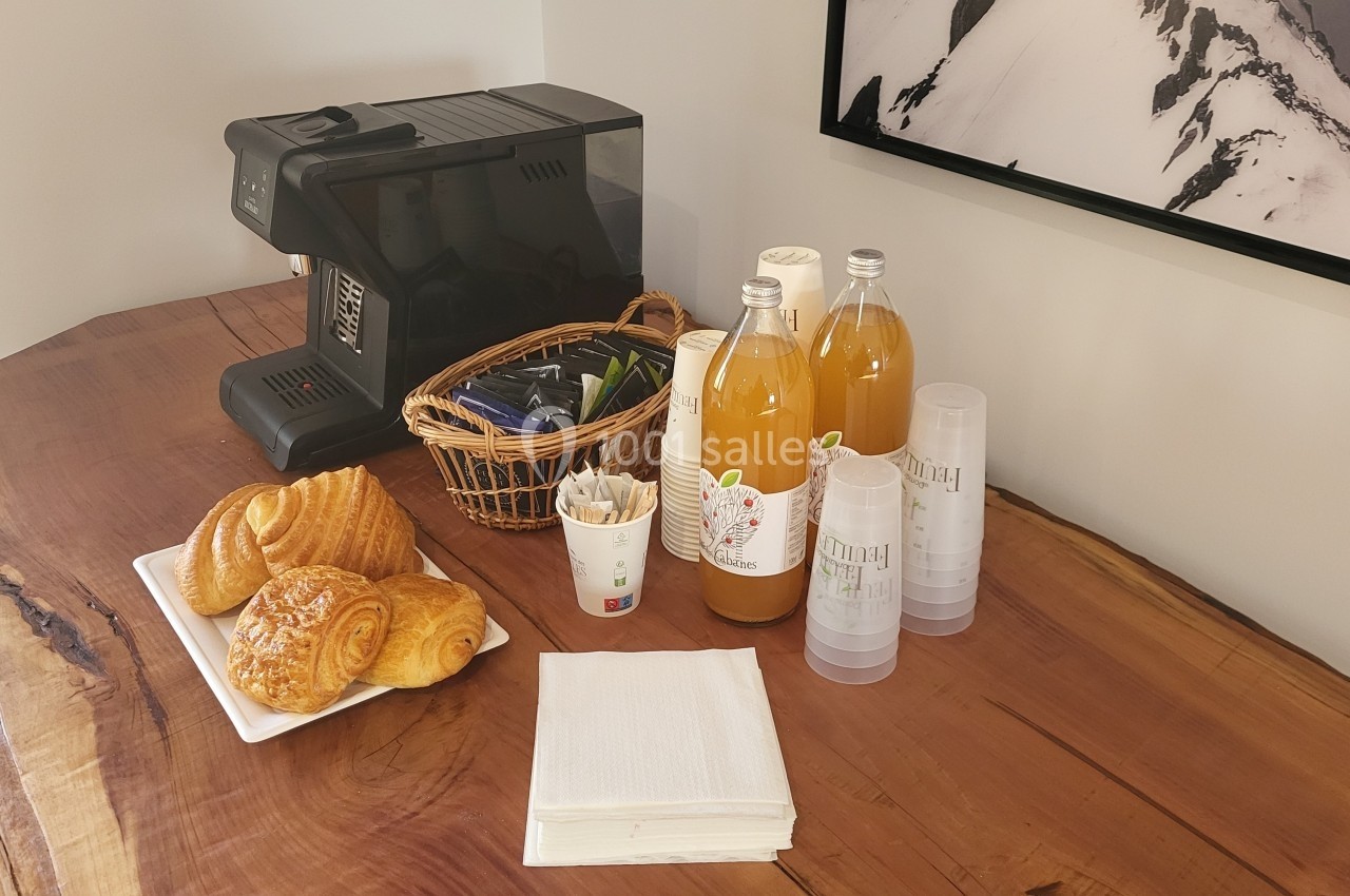 Petit-déjeuner avec viennoiseries, machine à café, jus de fruits en bouteilles et gobelets sur une table en bois.