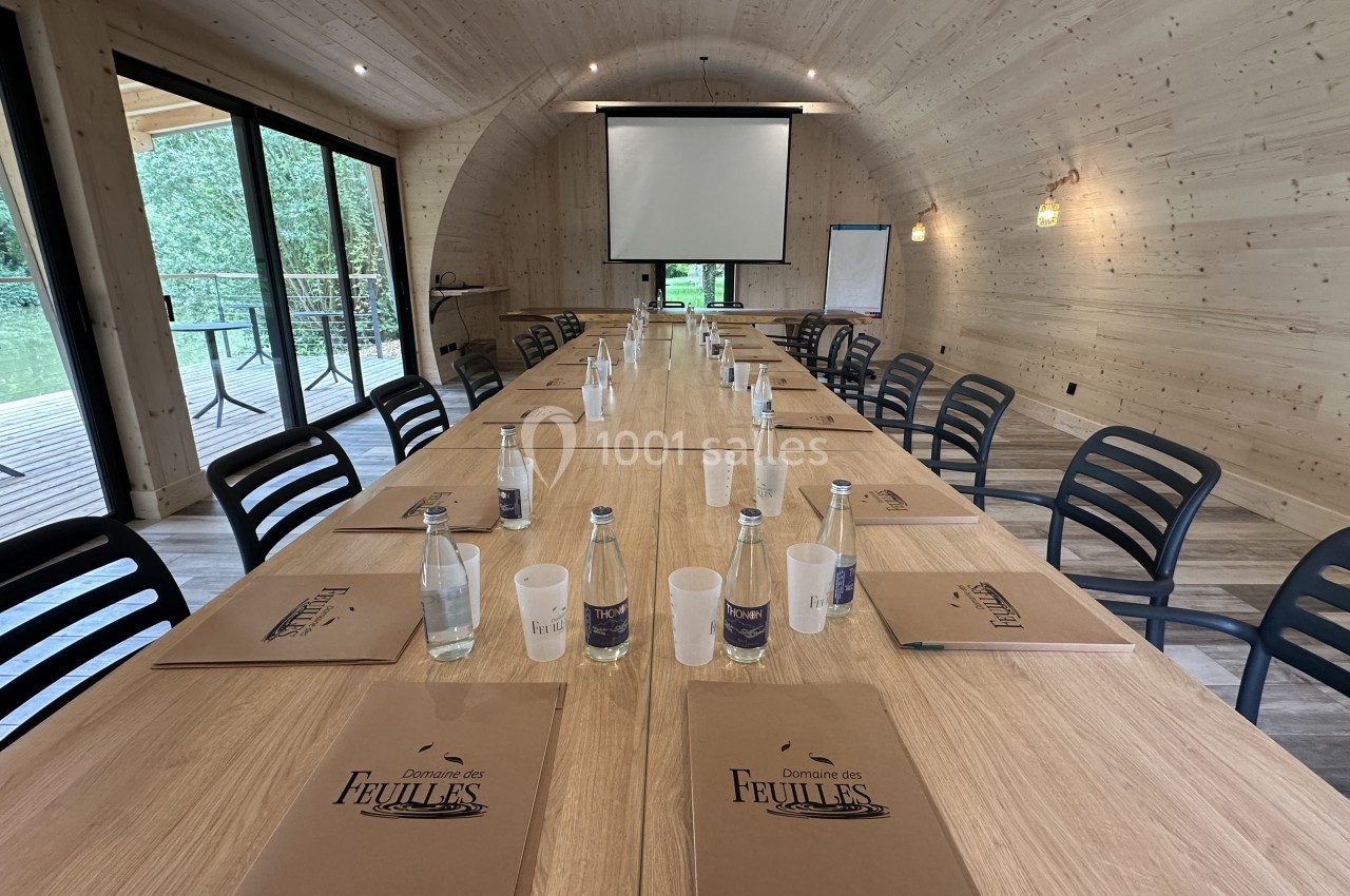 Salle de réunion en bois avec une grande table rectangulaire, chaises noires, écran de projection et vue sur l'extérieur.