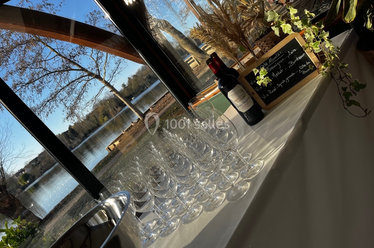 Verres à vin alignés sur une table près d'une fenêtre avec vue sur un lac, bouteille de vin et ardoise décorative.