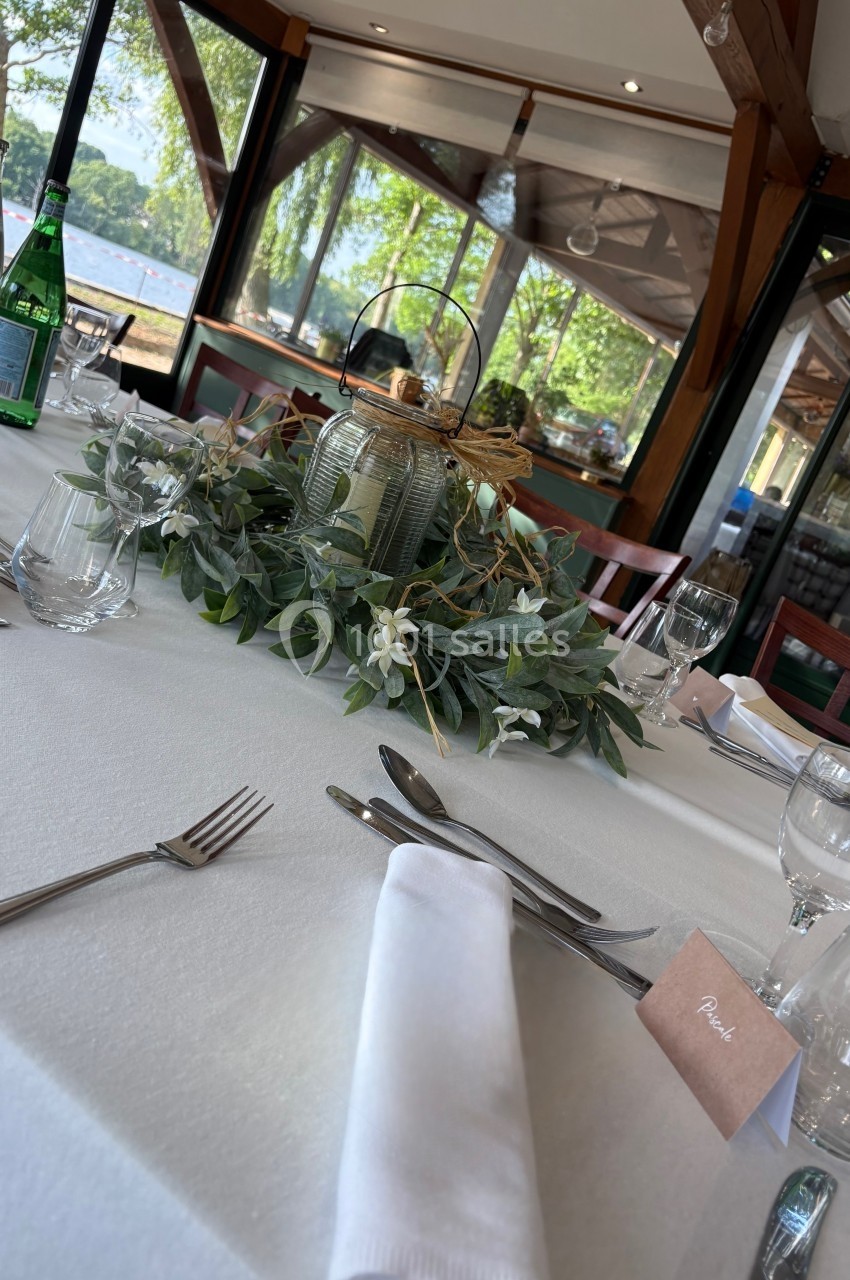 Table dressée avec une nappe blanche, couverts, verres, centre de table floral et vue sur un paysage extérieur verdoyant.
