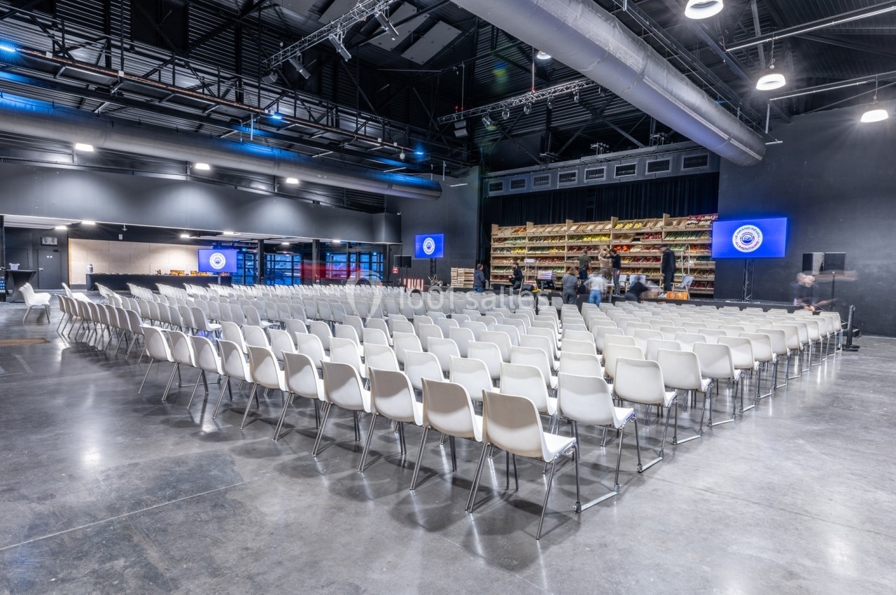 Salle de conférence moderne avec rangées de chaises blanches alignées, scène et écrans allumés en arrière-plan.