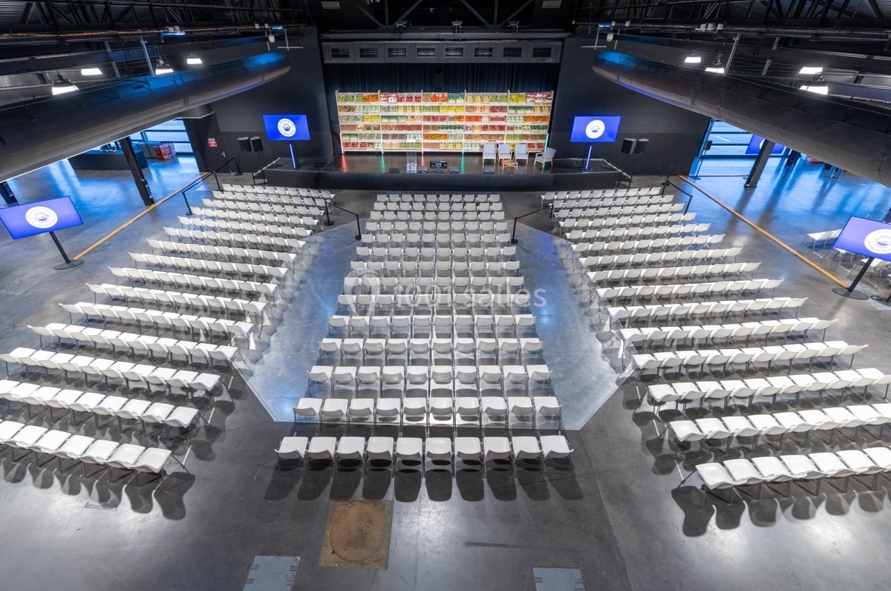 Salle de conférence avec des rangées de chaises blanches face à une scène équipée d'étagères colorées et d'écrans.