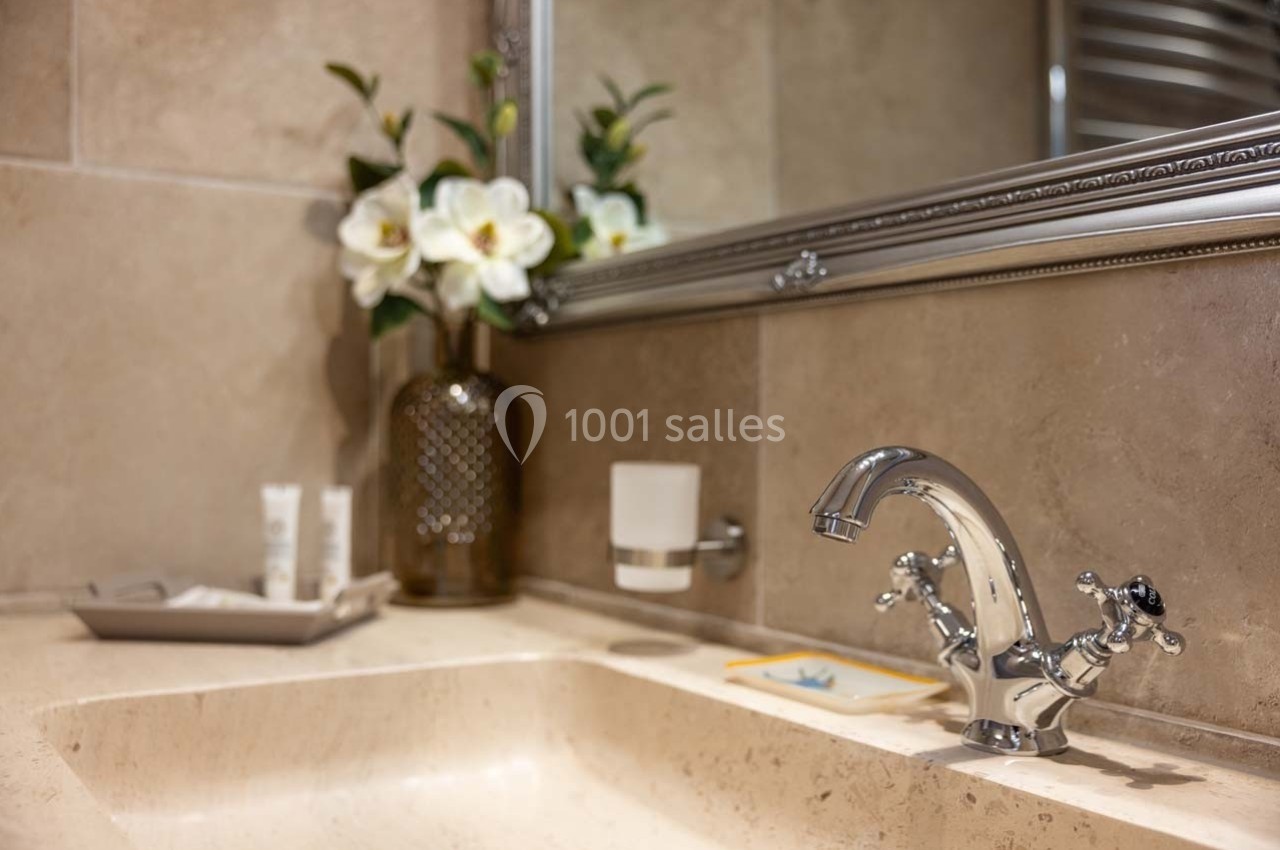 Lavabo en marbre beige avec un robinet chromé, un miroir encadré et un vase contenant des fleurs blanches.