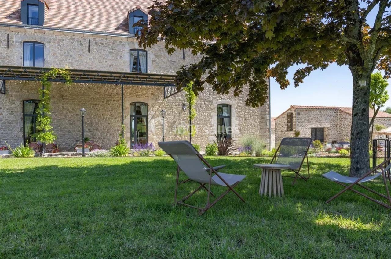 Chaises longues et table basse sur une pelouse ombragée devant un bâtiment en pierre avec des fenêtres et un arbre.