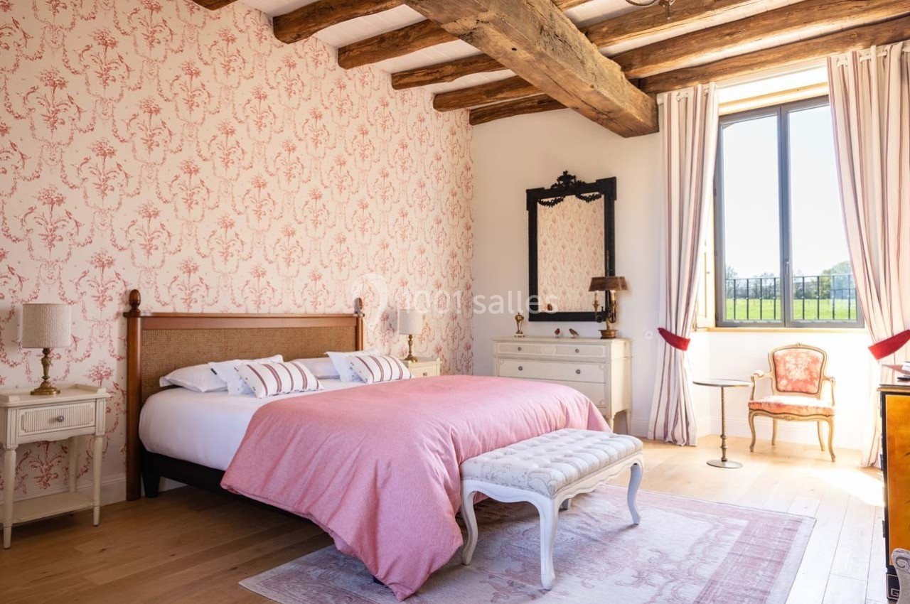 Chambre lumineuse avec lit double, murs ornés de papier peint floral, poutres apparentes et vue sur un jardin.